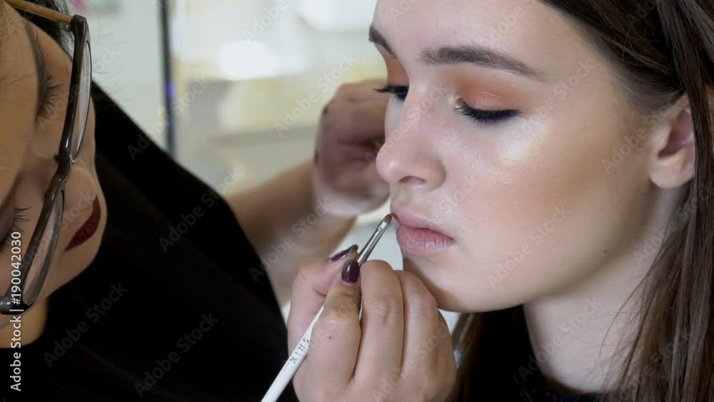 Asian female visage artist is applying lipstick to the lips of her young client. Model with beautiful make up is sitting straight while the lady master is putting color by the white brush in her hand Asian female visage artist is applying lipstick to the lips of her young client. Model with beautiful make up is sitting straight while the lady master is putting color by the white brush in her hand