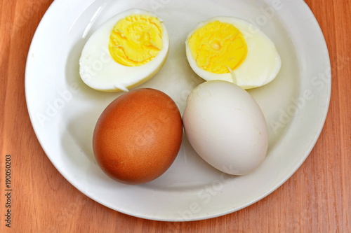 Half hard boiled chicken egg and whole red and white egg in eggshell on a white saucer and wooden table