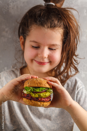 Beautiful happy hungry baby girl eating vegan burger.  Vegan beet chickpea burgers with vegetables, guacamole and rye buns. Healthy child vegan food concept.