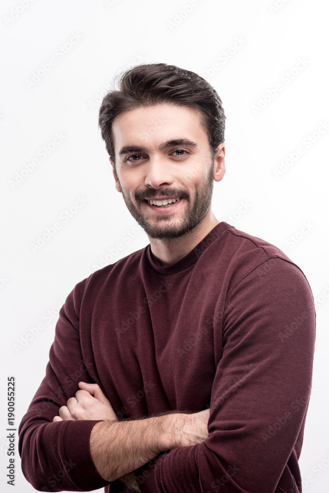 Pleasant smile. Handsome bristled young man in a burgundy sweater posing against a white background and folding his arms across his chest while smiling at the camera Pleasant smile. Handsome bristled young man in a burgundy sweater posing against a white background and folding his arms across his chest while smiling at the camera