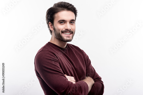 Very masculine. Cheerful handsome man standing half-turned and folding his arms across his chest while posing against a white background