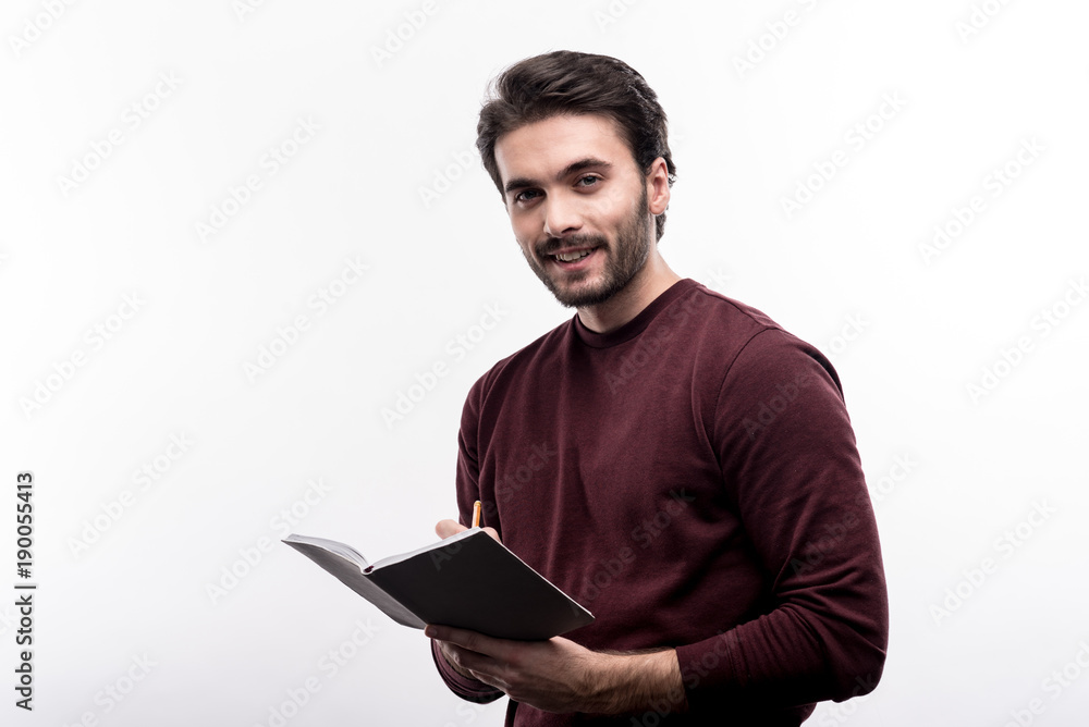 Important remarks. Charming young man taking notes in his daily planner and smiling at the camera while posing against a white background Important remarks. Charming young man taking notes in his daily planner and smiling at the camera while posing against a white background