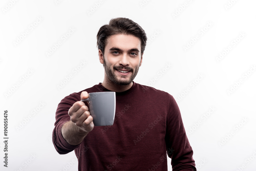 Treat yourself. Handsome dark-haired young man in a burgundy sweater offering a cup of coffee while posing against a white background Treat yourself. Handsome dark-haired young man in a burgundy sweater offering a cup of coffee while posing against a white background