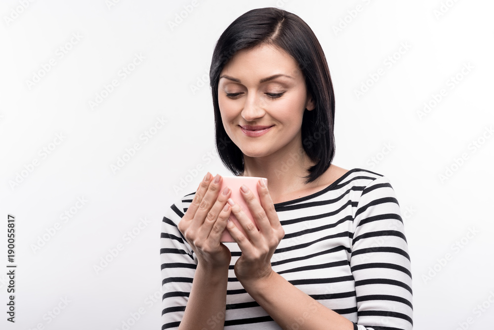 Pleasant aroma. Beautiful dark-haired young woman cupping the mug of coffee with her both hands and smiling while standing isolated on a white background Pleasant aroma. Beautiful dark-haired young woman cupping the mug of coffee with her both hands and smiling while standing isolated on a white background