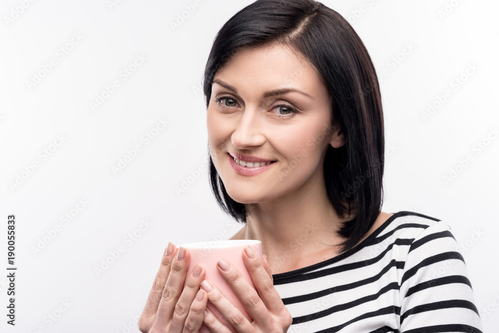 Warm drink. Pleasant pretty woman in a striped pullover holding a pink mug of coffee with her both hands and posing for the camera while smiling Warm drink. Pleasant pretty woman in a striped pullover holding a pink mug of coffee with her both hands and posing for the camera while smiling