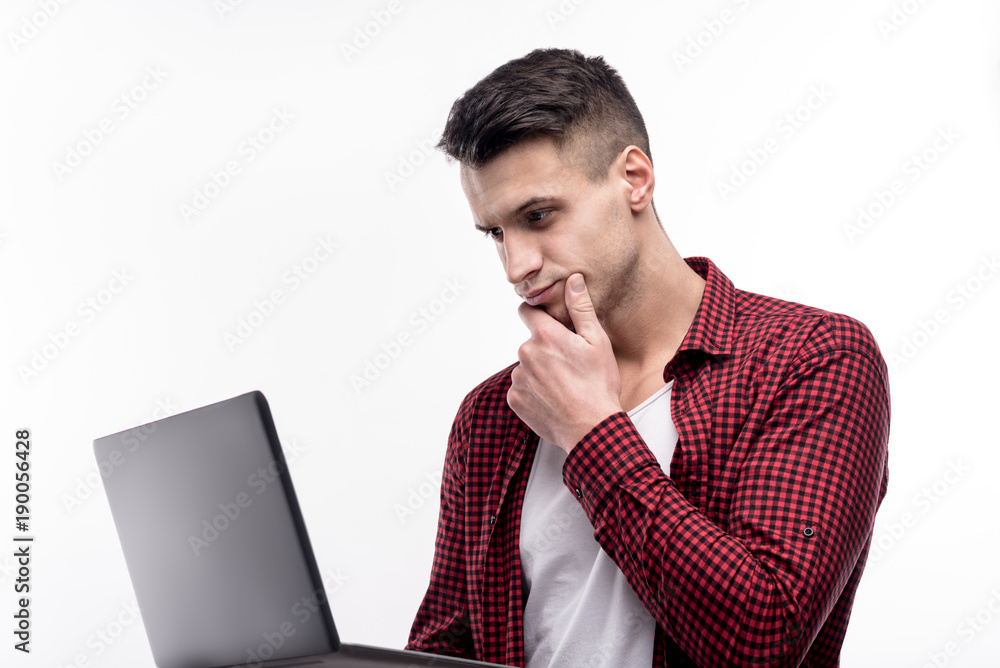Interesting content. Pleasant young man holding a laptop and reading from it while resting his chin on his hand and paying all attention to the text Interesting content. Pleasant young man holding a laptop and reading from it while resting his chin on his hand and paying all attention to the text