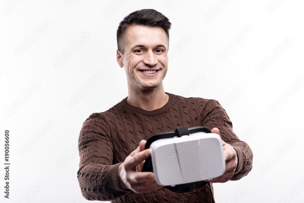 Best purchase. Pleasant handsome man in a brown sweater showing his new virtual reality headset and smiling contently Best purchase. Pleasant handsome man in a brown sweater showing his new virtual reality headset and smiling contently