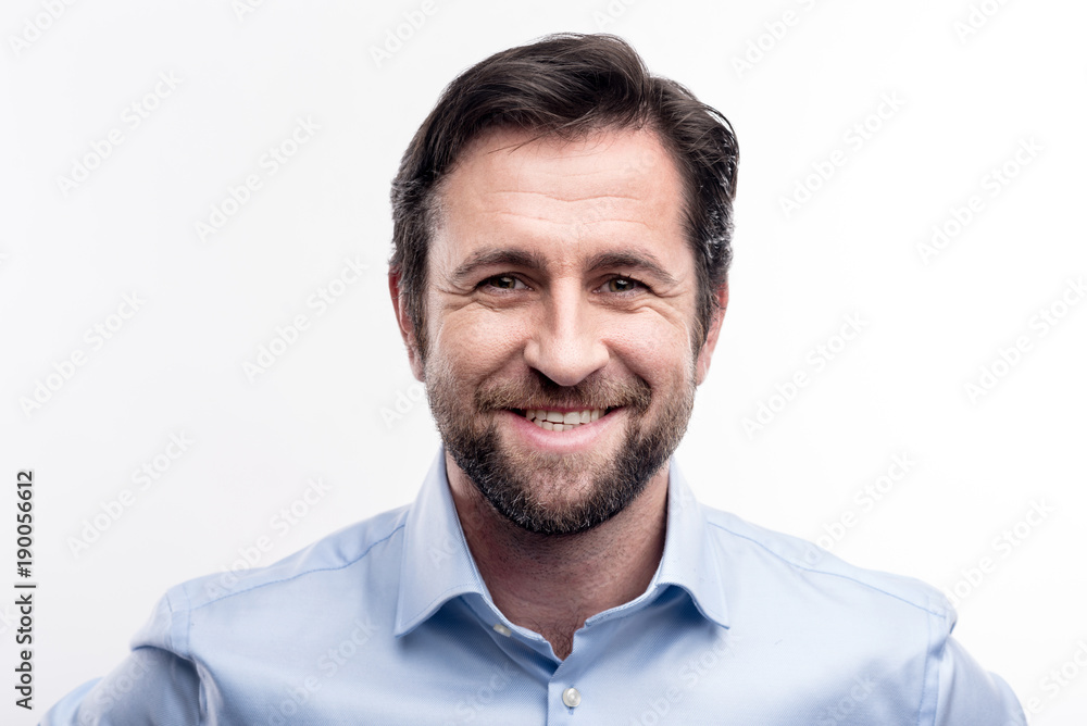 Appealing smile. The portrait of a handsome dark-haired middle-aged man in a blue shirt smiling at the camera while posing against a white background Appealing smile. The portrait of a handsome dark-haired middle-aged man in a blue shirt smiling at the camera while posing against a white background