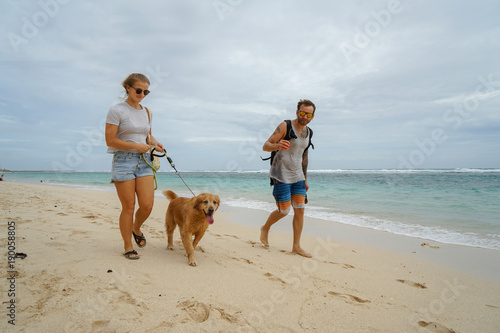 A family of freelancers with a dog is walking along the beach. A stylish young girl leads a gold retriever next to her husband with a surfer. Close-up.
