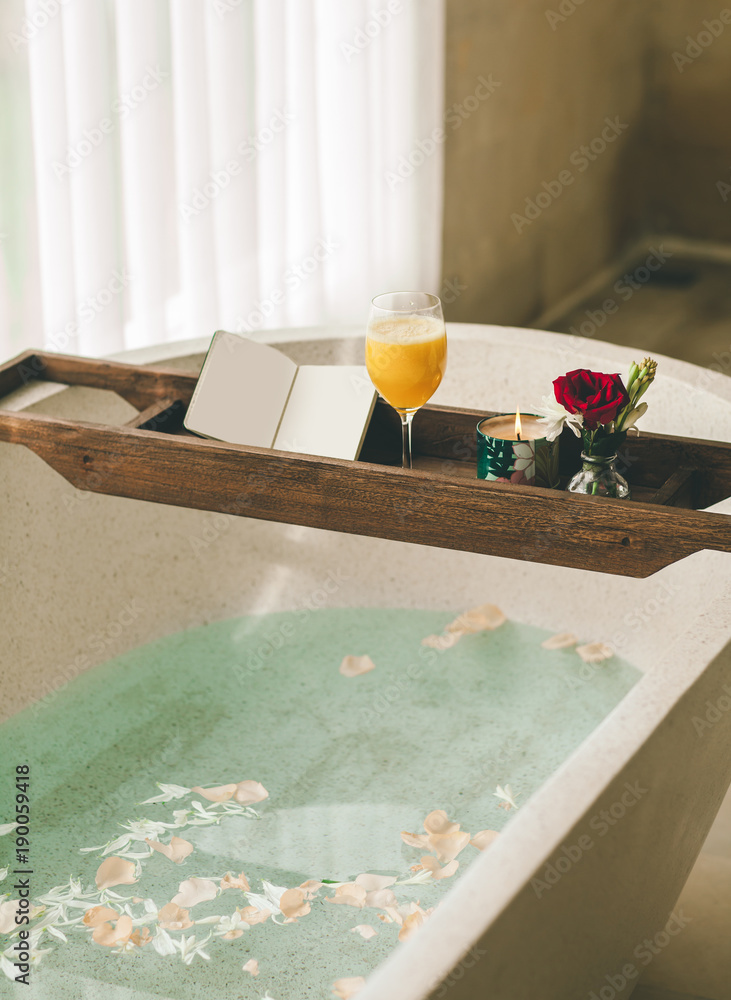 Bright Bathroom at Home. Bath with Flower Petals and Salt. Tray over the Bath with Orange Juice, Notepad and Candles.