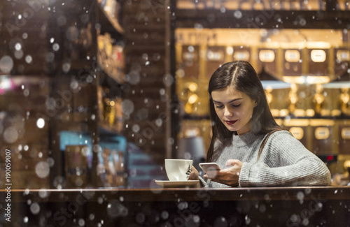 Young pretty brunette girl wears grey sweater holding smartphone sitting near cafe window in snowy Christmas time