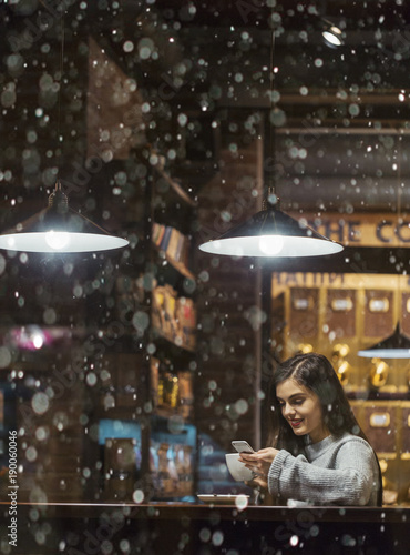 Young pretty brunette girl wears grey sweater holding smartphone sitting near cafe window in snowy Christmas time