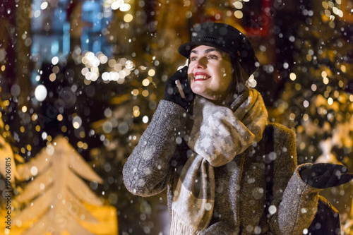 Stylish brunette smiling girl having pleasant talk by smartphone before garland wall in city snowy evening Christmas time