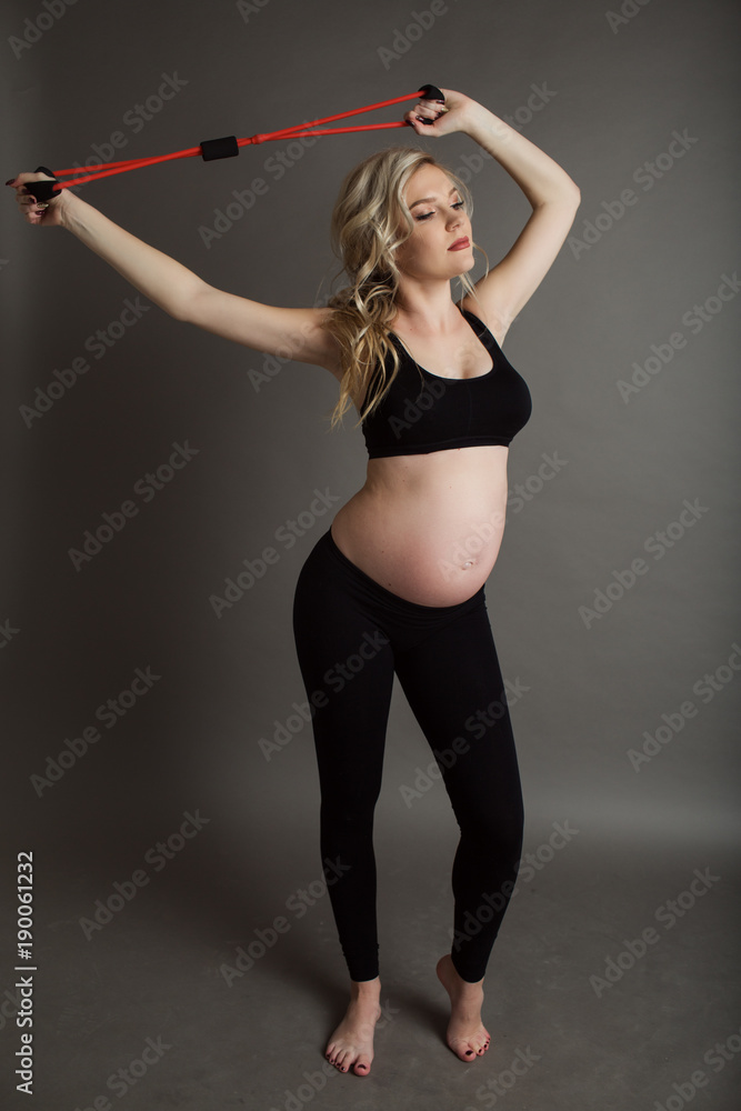 Pregnant beautiful blonde woman is wearing sportive black clothes doing exercises with rubber band isolated on gray, healthy life concept