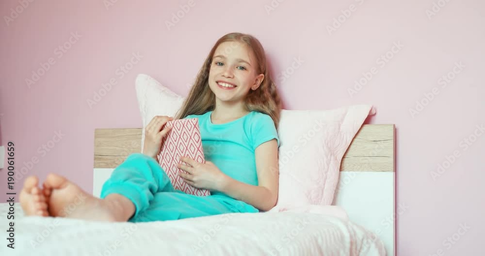Schoolgirl holds book and opens it sitting on bed and smiling Schoolgirl holds book and opens it sitting on bed and smiling