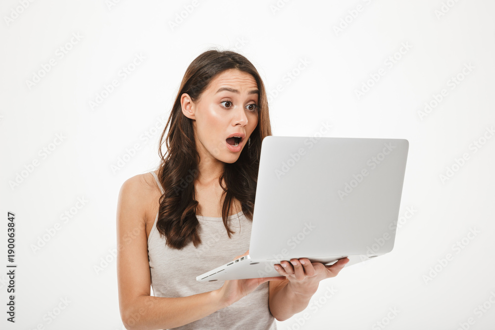 Photo of surprised brunette woman looking on screen of new notebook being excited or thrilled, isolated over white wall Photo of surprised brunette woman looking on screen of new notebook being excited or thrilled, isolated over white wall
