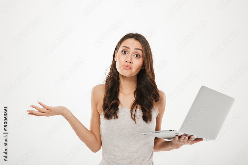 Photo of upset woman with long brown hair throwing up hands and expressing confusion while holding silver personal computer, isolated over white wall Photo of upset woman with long brown hair throwing up hands and expressing confusion while holding silver personal computer, isolated over white wall