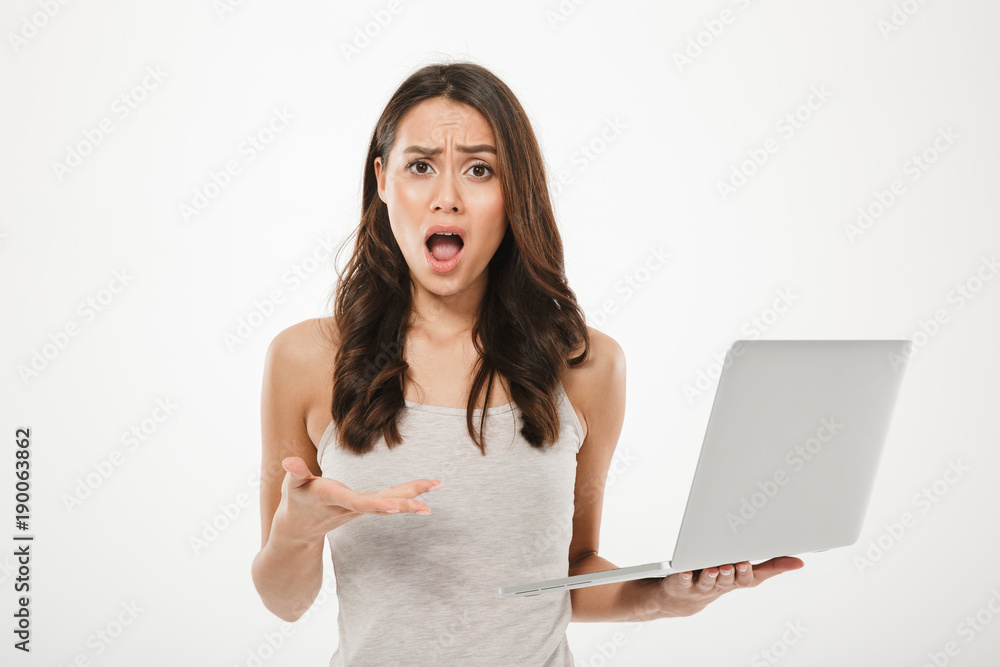 Photo of brunette woman looking on camera in perplexion and holding silver notebook in hand, isolated over white wall Photo of brunette woman looking on camera in perplexion and holding silver notebook in hand, isolated over white wall