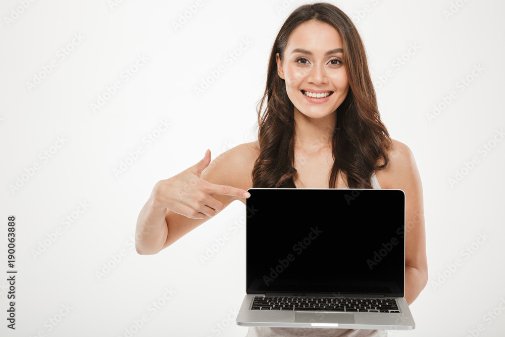 Horizontal photo of happy woman 30s smiling and demonstrating black empty screen of silver laptop on camera with finger, over white wall Horizontal photo of happy woman 30s smiling and demonstrating black empty screen of silver laptop on camera with finger, over white wall