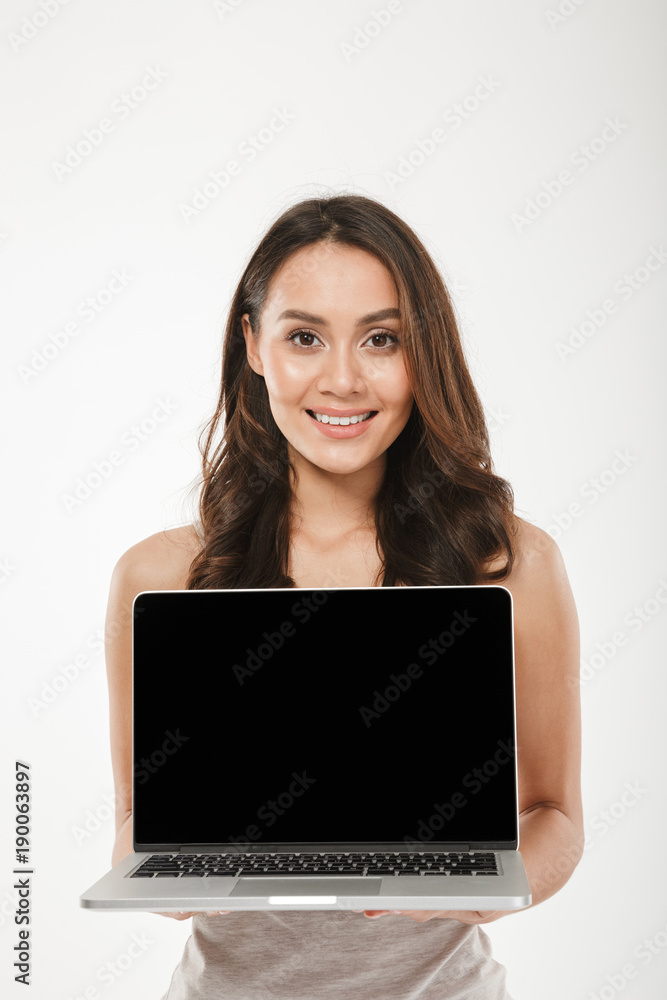 Vertical image of adult woman 30s smiling and showing black empty screen of silver laptop on camera, over white wall Vertical image of adult woman 30s smiling and showing black empty screen of silver laptop on camera, over white wall