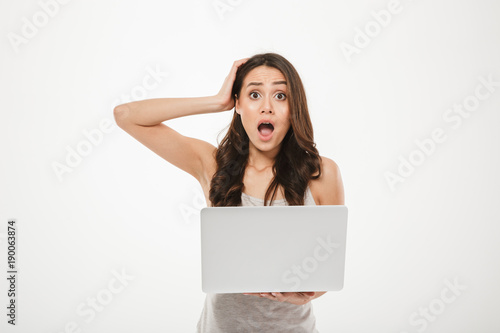 Photo of uptight woman 30s looking on camera and grabbing her head with open mouth while using silver notebook, isolated over white wall