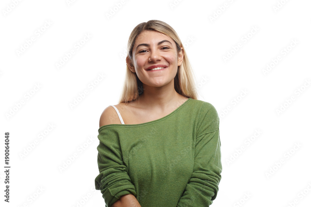 Horizontal shot of positive young European female student with nose ring and dyed hair posing in studio, looking at camera with happy charming smile, feeling relaxed after lectures at university
