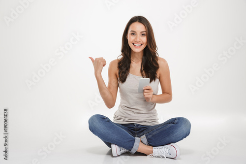 Photo of pleased woman with long brown hair sitting with legs crossed on the floor using silver smartphone and pointing away, isolated over white wall