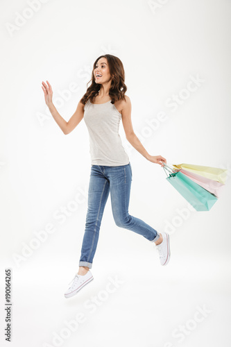 Full-length image of ecstatic adult female levitating or jumping with lots of colorful shopping bags in hand, isolated over white background