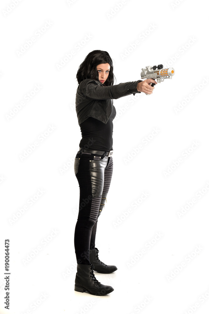 full length portrait of black haired girl wearing leather outfit. standing pose while holding a gun, isolated on a white studio background. full length portrait of black haired girl wearing leather outfit. standing pose while holding a gun, isolated on a white studio background.