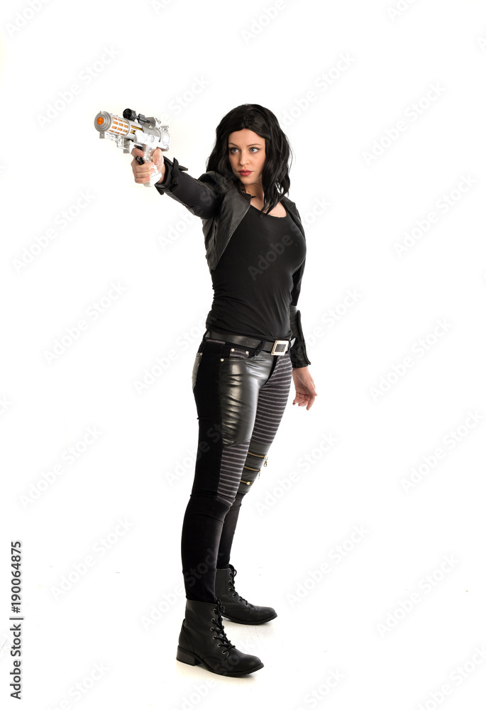full length portrait of black haired girl wearing leather outfit. standing pose while holding a gun, isolated on a white studio background. full length portrait of black haired girl wearing leather outfit. standing pose while holding a gun, isolated on a white studio background.