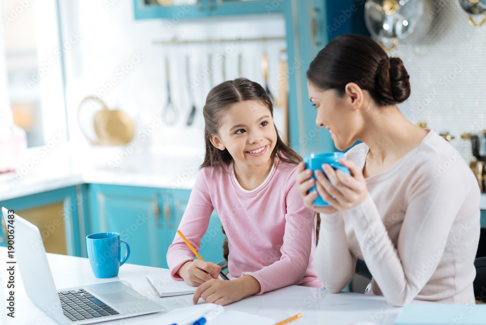 My smart child. Pretty alert dark-haired schoolgirl writing in her notebook and smiling at her mother standing near the girl with a cup of tea