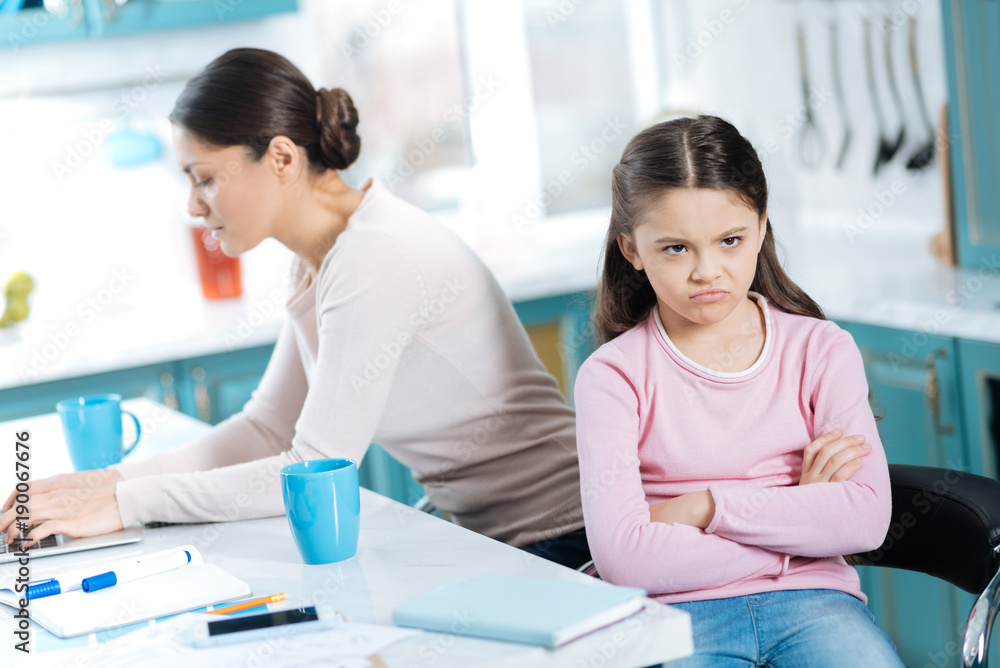 Fototapeta premium Boredom. Sweet dissatisfied dark-eyed girl having her arms crossed and sitting near her mom working on her laptop