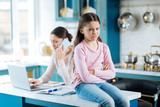 Unhappy. Pretty miffed dark-eyed girl holding her arms crossed and sitting near her mom working on her laptop
