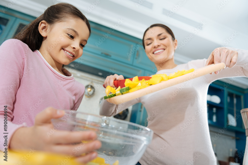 Cooking process. Pretty inspired dark-haired mother smiling and putting ingredients into a bowl and making a salad with her daughter Cooking process. Pretty inspired dark-haired mother smiling and putting ingredients into a bowl and making a salad with her daughter