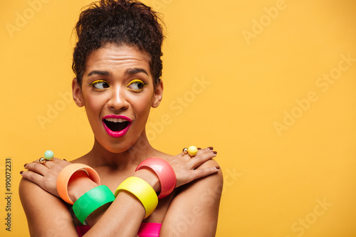 Gorgeous afro american woman in colorful adornment embarrassing while posing on camera half-naked with crossed hands on shoulders, over yellow copy space