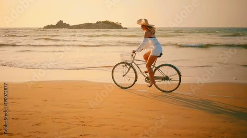 Woman in white and hat mooving on her bicycle along beach sand at summer time under burning sunset sky, lifestyle carefree.