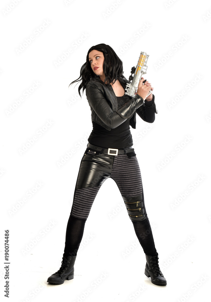 full length portrait of black haired girl wearing leather outfit. standing pose while holding a gun, isolated on a white studio background. full length portrait of black haired girl wearing leather outfit. standing pose while holding a gun, isolated on a white studio background.