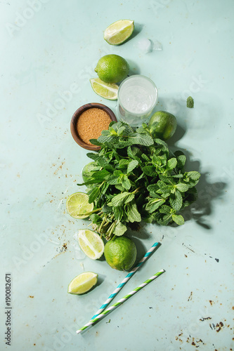 Ingredients for making mojito cocktail. Bundle of fresh mint, whole and sliced limes, brown sugar, crashed ice cubes, glass of soda water, cocktail tubes over green pin up background. Top view, space