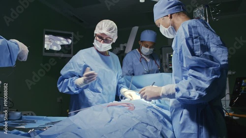 PAN of surgical team in scrubs and masks operating on patient and stitching incision during procedure in sterile room photo