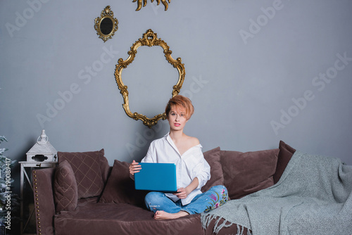 Stylish young woman with laptop in hands on sofa and looking at camera . Dressed in a white shirt and jeans with embroidery. Gray wall with shadow behind her. photo