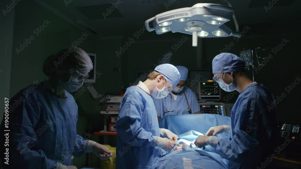 Zoom out shot of team of surgeons wearing scrubs and masks performing procedure on patient in sterile room with modern equipment Zoom out shot of team of surgeons wearing scrubs and masks performing procedure on patient in sterile room with modern equipment