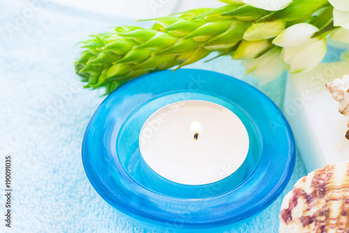 Aromatherapy Spa Concept with a fragrant candle in a blue glass candle holder  sea shells and white flower on white wooden background  close up