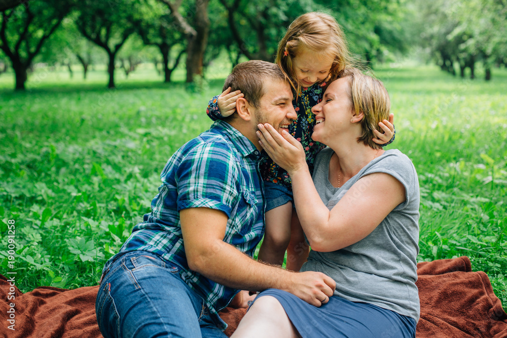 Fototapeta premium Young happy family of three lying on blanket in the park having fun. Happy parenting concept. Little girl with mother and father outdoors