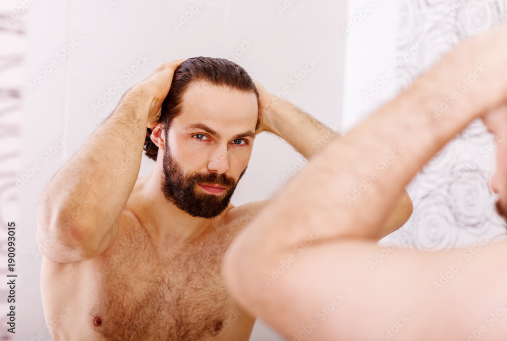 Handsome young man touching his hair with hand and smiling while standing in front of the mirror Handsome young man touching his hair with hand and smiling while standing in front of the mirror