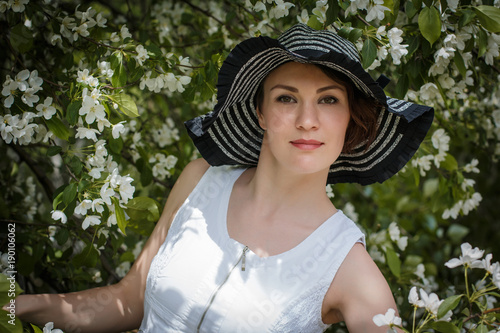 Nice girl in white dress with her black hat and apple tree with white flowers behind