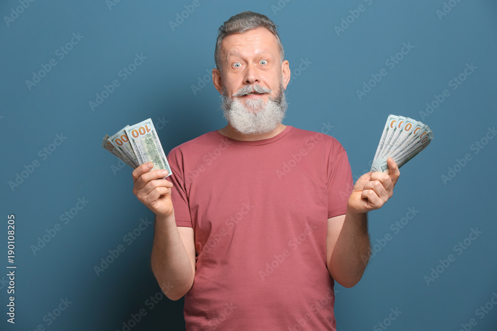 Emotional senior man with lot of dollar banknotes on color background