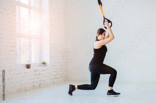 Beautiful brunette woman in black sportswear training with suspension trainer sling on white loft interior background. photo