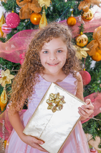 Beautiful smiling curly girl wearing a pink dress and holding a huge gift in her hands, with a Christmas tree behind, christmas and new year concept