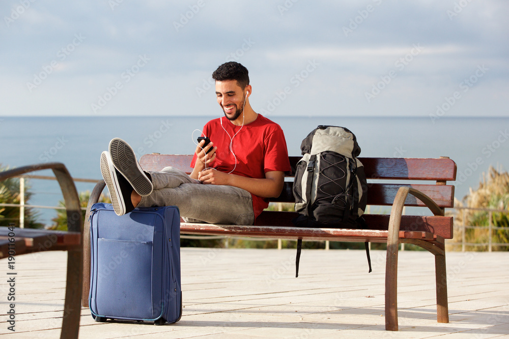 Fototapeta premium happy young man sitting on bench with luggage and listening to music with mobile phone and earphones