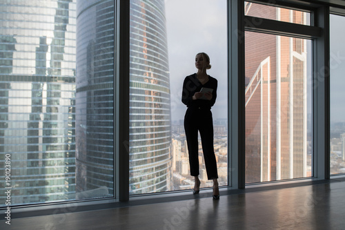 Business woman holding a tablet and standing in a modern office. Panoramic windows background.